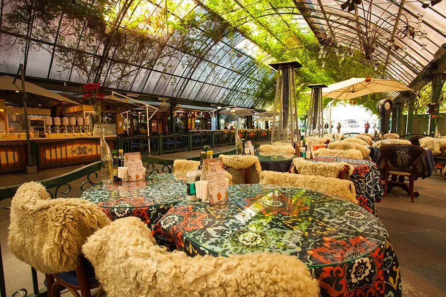 Área de café ao ar livre sob um toldo de vidro com toalhas de mesa estampadas e cadeiras cobertas com mantas de pele sintética cor creme.