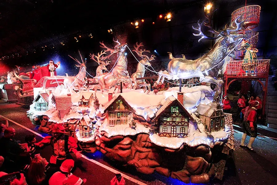 Um carro alegórico iluminado de vários andares apresenta Papai Noel, renas e casas em miniatura cobertas de neve à noite.