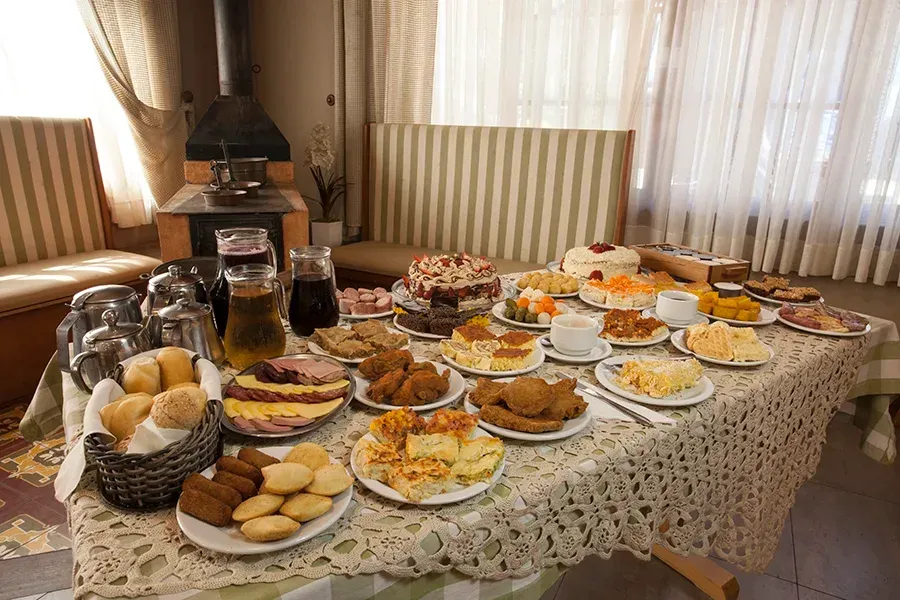 Uma mesa posta com diversos pães, doces, carnes e bebidas em uma sala de jantar aconchegante com cortinas brancas.