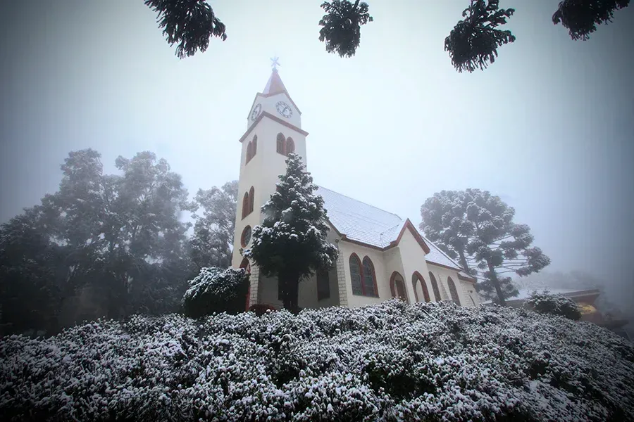 Uma igreja cor creme com uma torre sineira ergue-se numa colina coberta de neve, rodeada por árvores e envolta num nevoeiro denso.