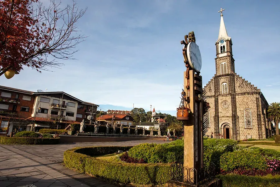 Uma torre do relógio ergue-se na praça da cidade, em frente a uma igreja de pedra com uma alta torre sineira, sob um céu azul límpido.