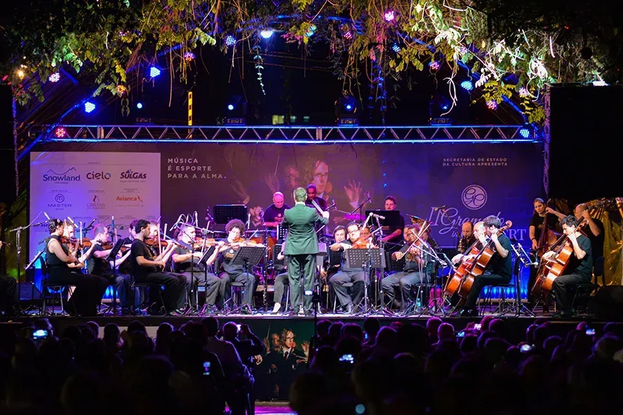 Uma orquestra se apresenta em um palco ao ar livre à noite, regida por um maestro que está de costas para o público.