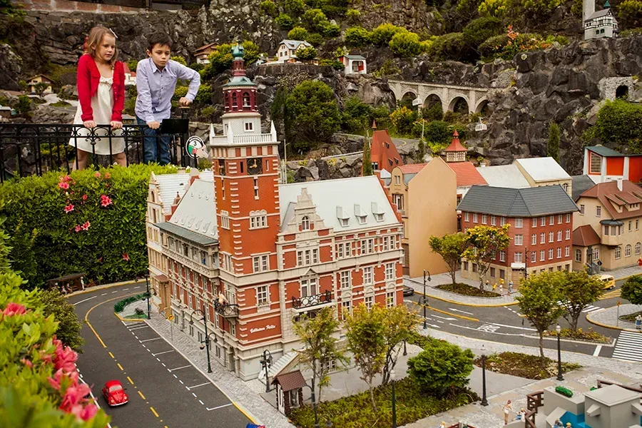 Duas crianças observam por cima de uma grade uma maquete detalhada de uma cidade em miniatura, que apresenta um imponente edifício de tijolos vermelhos com uma torre.