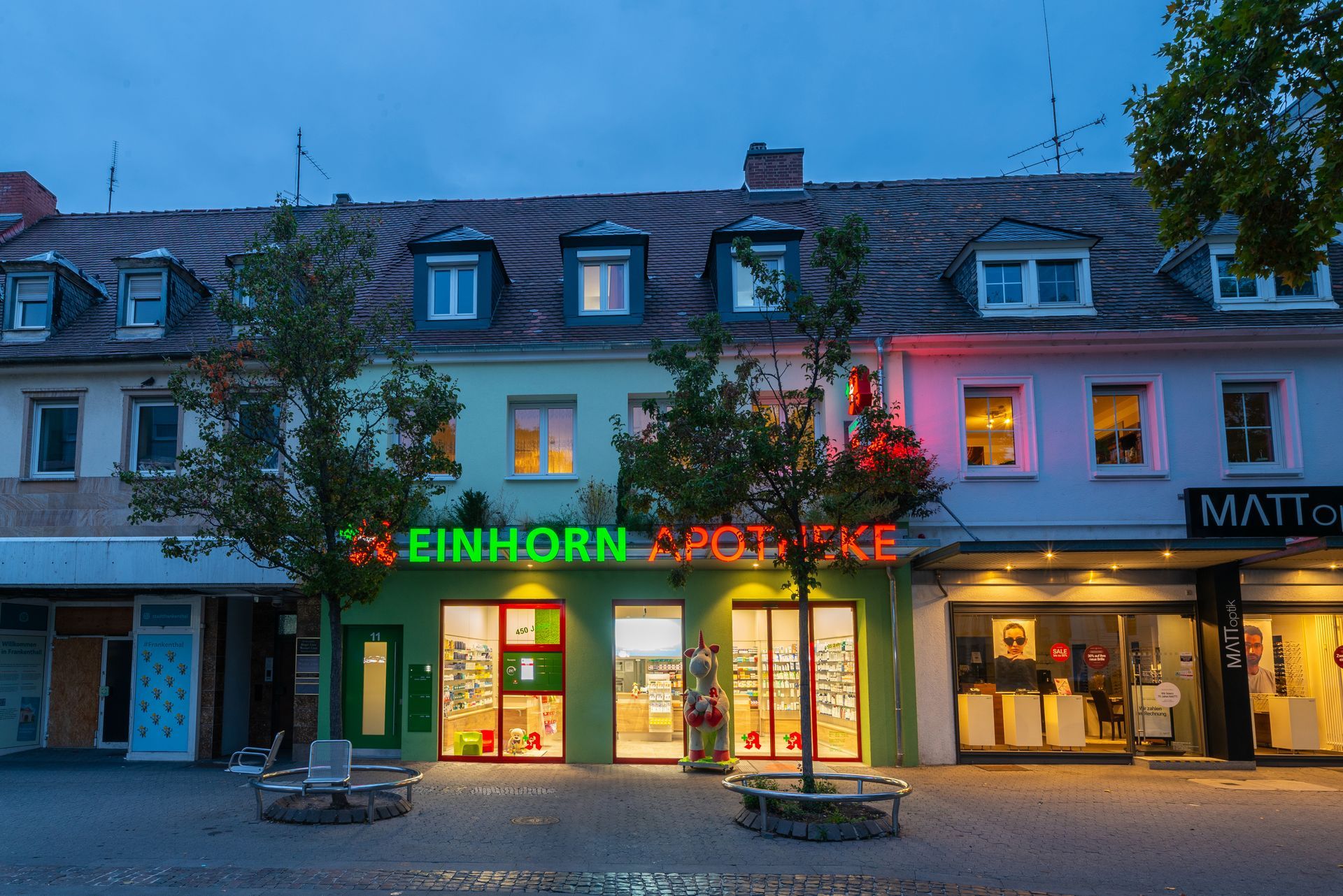 Außenansicht der Einhorn Apotheke mit Leuchtreklame an einer Straße in der Abenddämmerung.