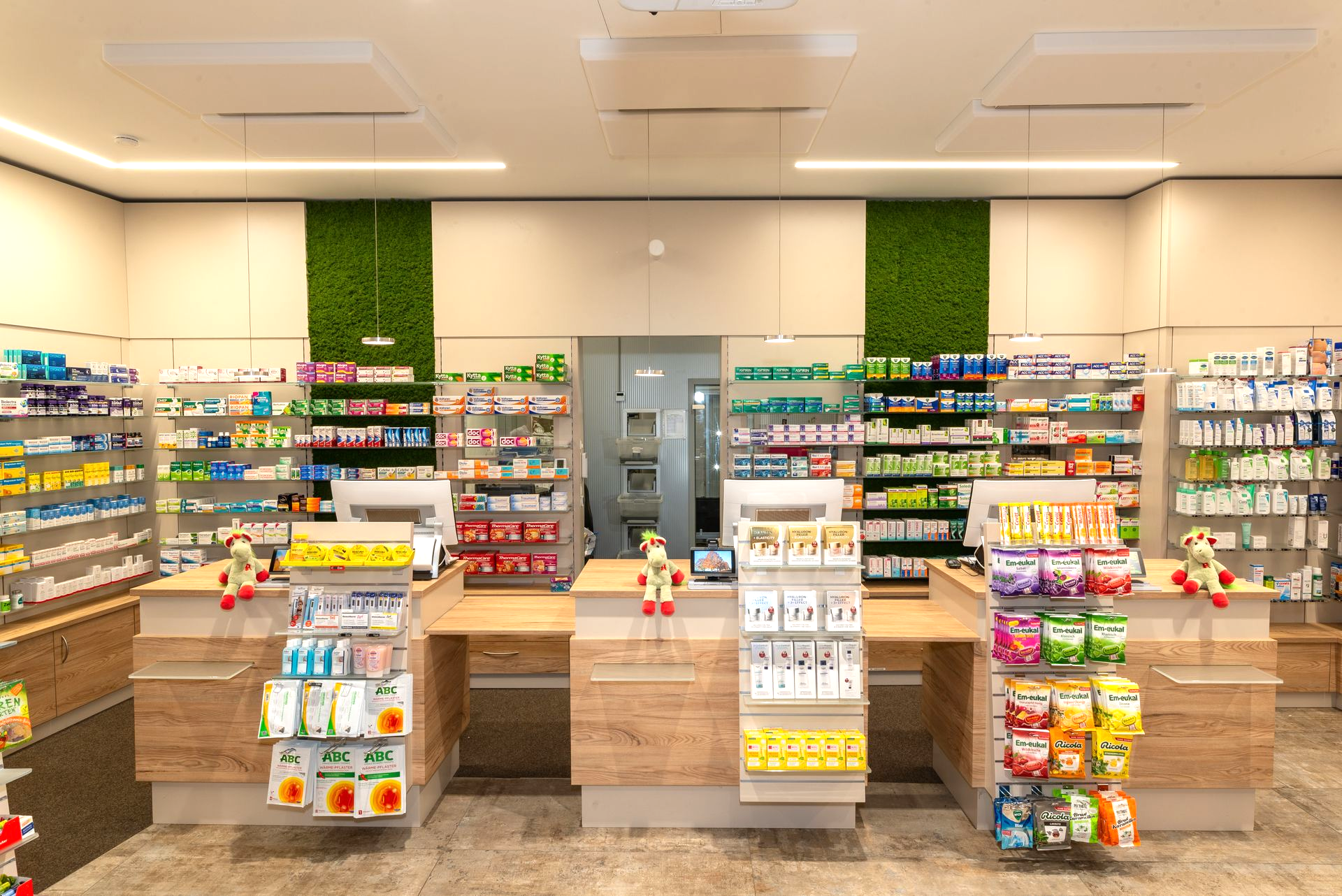 Innenansicht einer Apotheke mit Regalen voller Produkte und mehreren Kassen. Grüne Wandakzente.