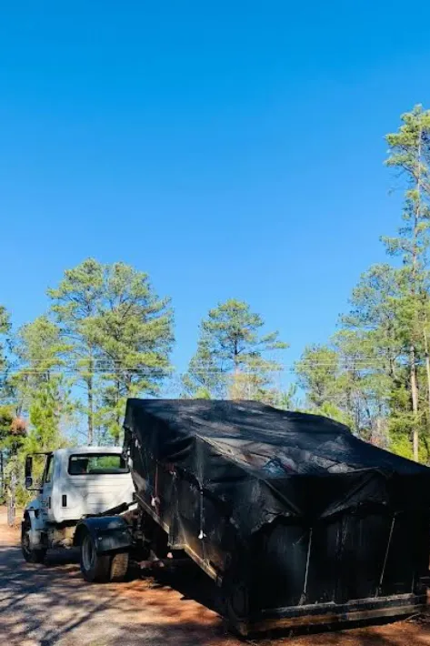 White truck towing a black tarp-covered trailer on a dirt road surrounded by tall pine trees and blue sky