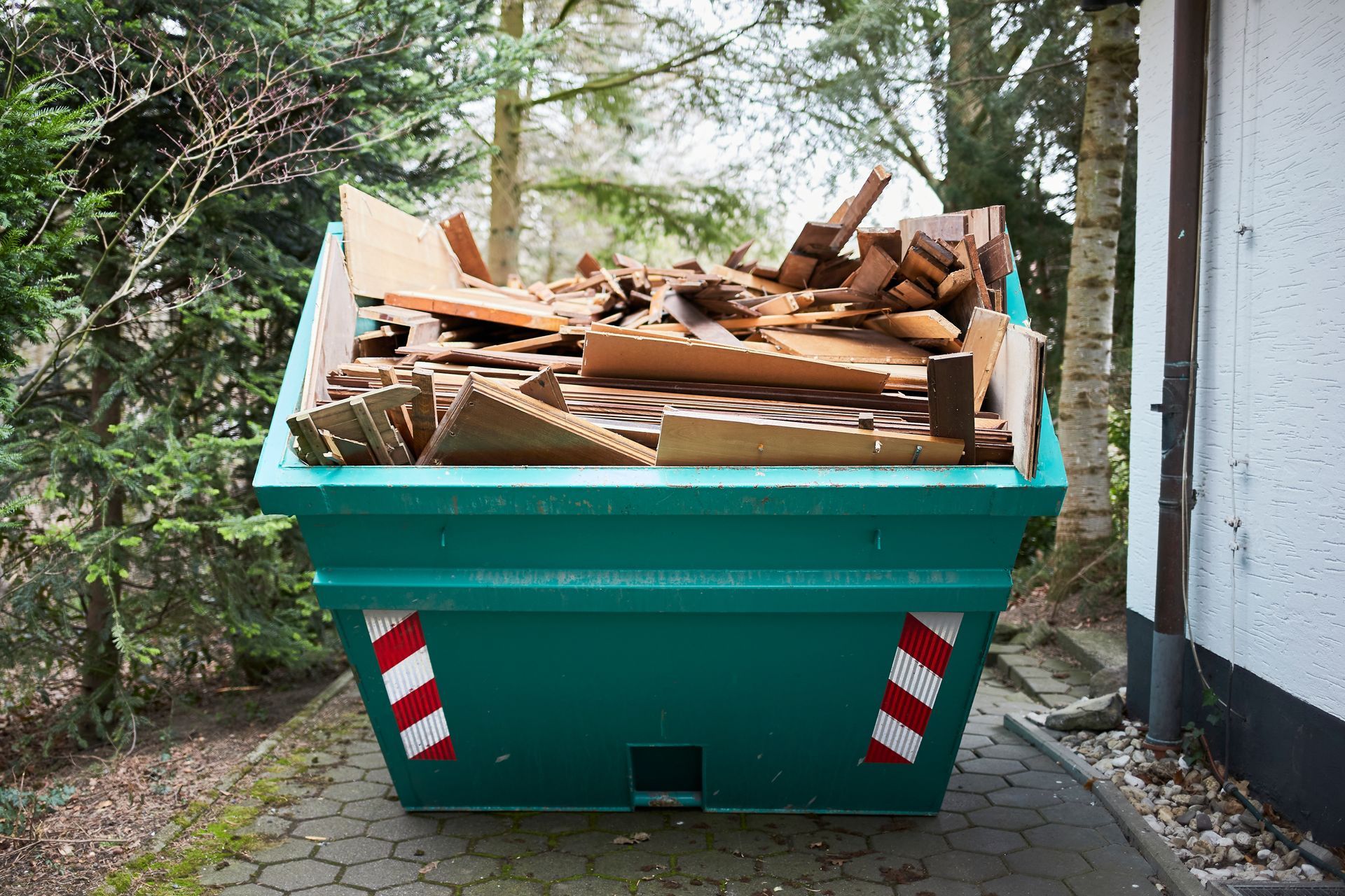 Green dumpster full of wood scraps and debris beside a building in a wooded area