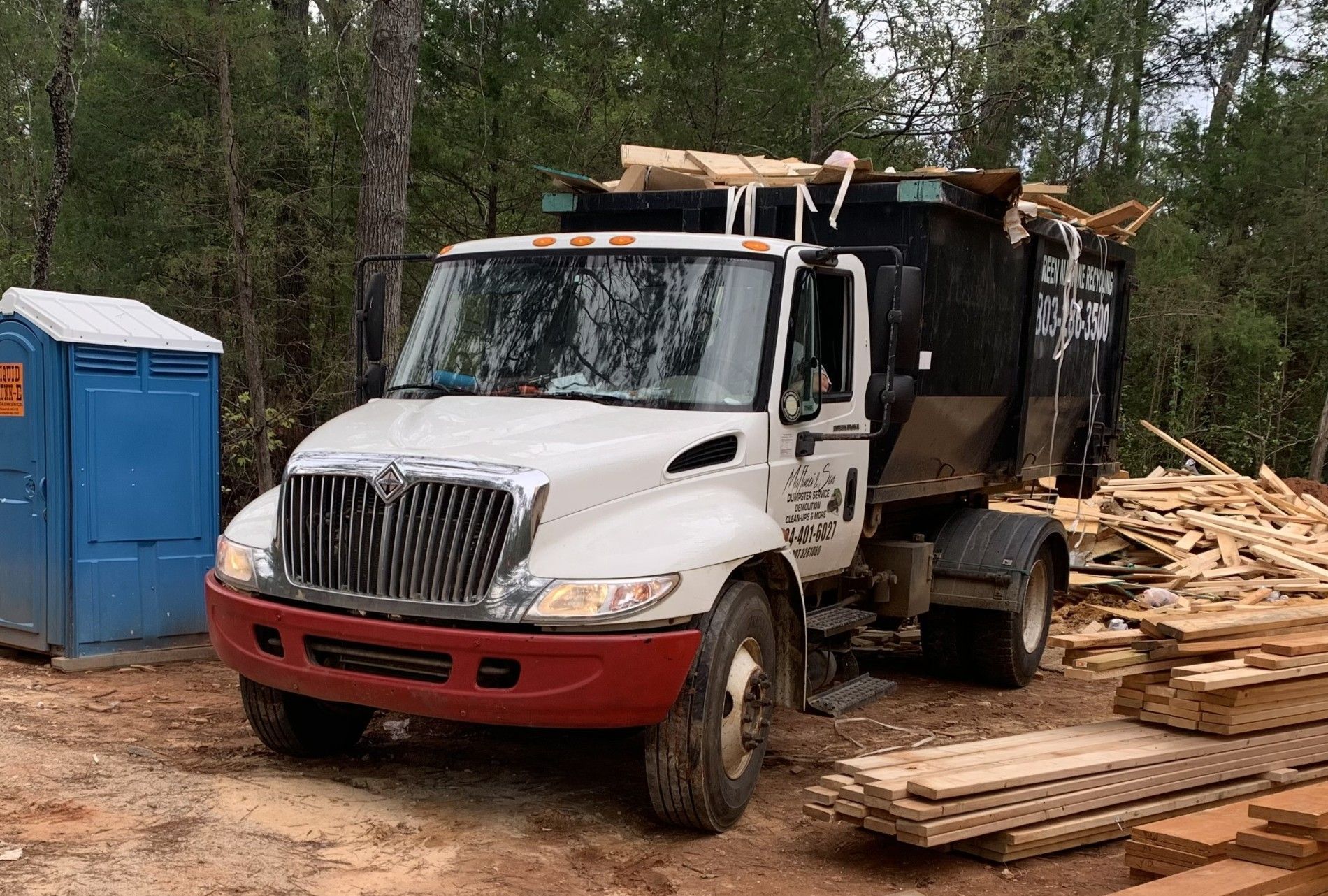 White dump truck at a wooded construction site beside blue portable toilets and piles of lumber