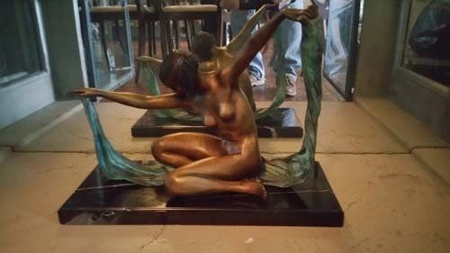 Bronze sculpture of a nude figure kneeling, arms outstretched, framed by green, vine-like structures, on a black marble base.