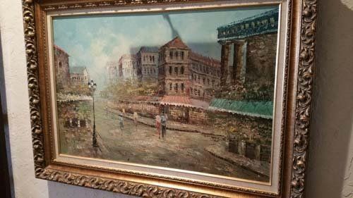 Oil painting of a European street scene with buildings, people, and lampposts, framed in gold.