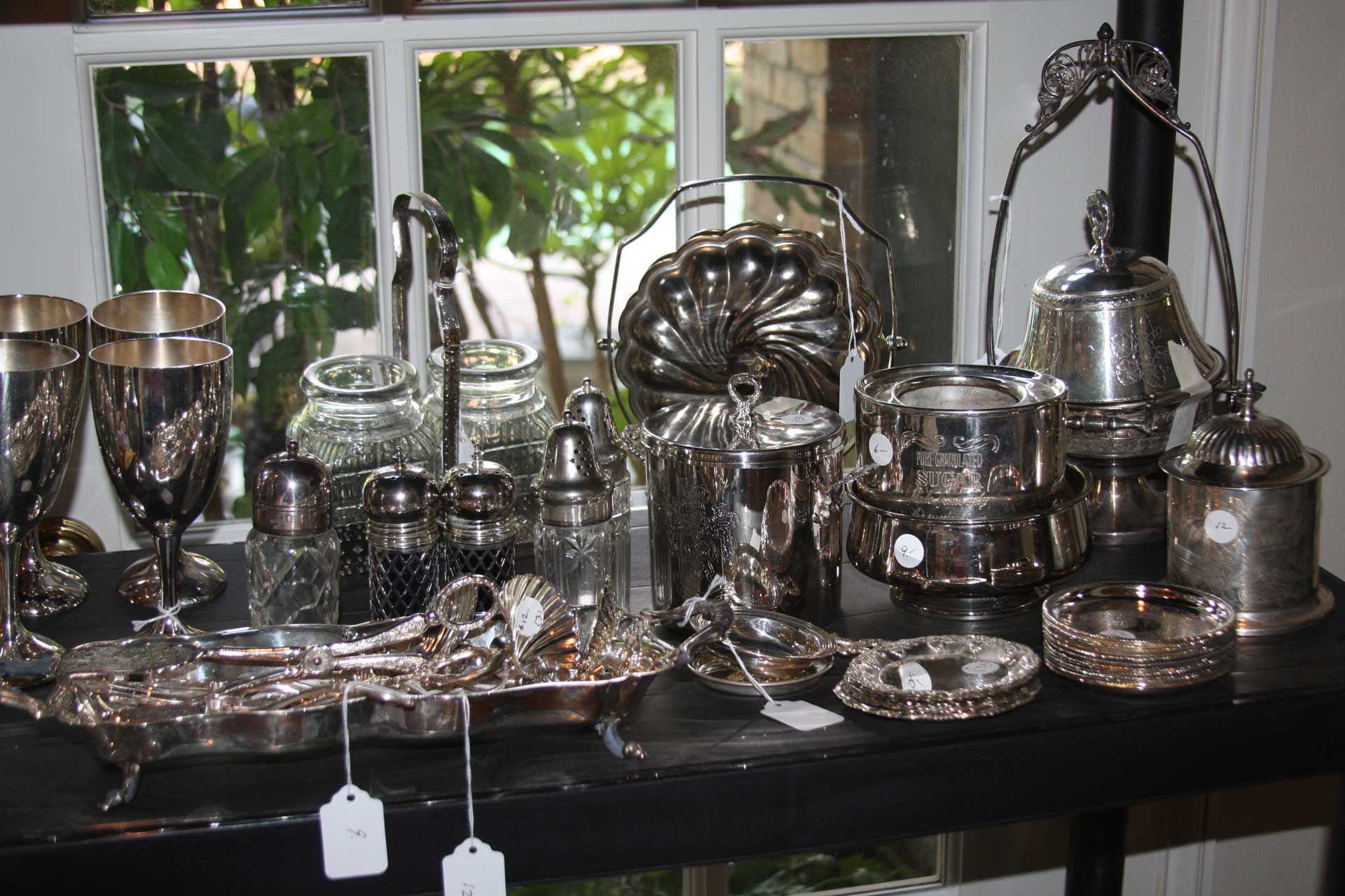 Silverware display: various silver serving pieces and accessories on a table near a window.