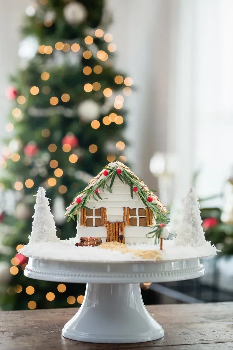 A gingerbread house is sitting on a cake stand in front of a christmas tree. A gingerbread house is sitting on a cake stand in front of a christmas tree.