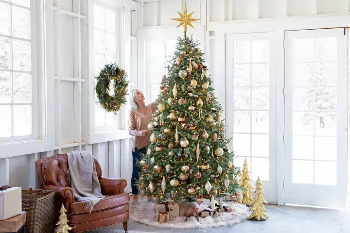 Woman decorates a Christmas tree in a bright, white-walled room. Tree features gold ornaments, lit with lights.