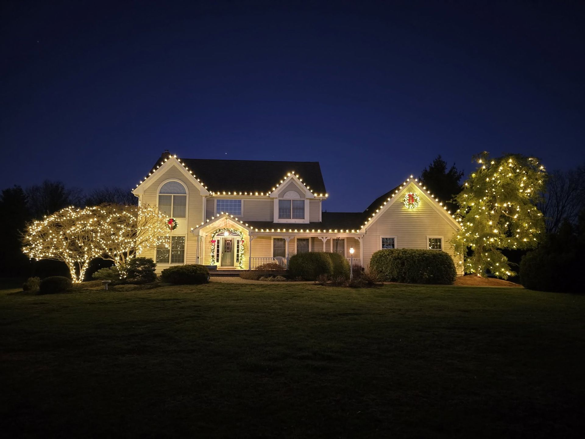 House at night outlined with white holiday lights, with lit trees and a dark lawn in front