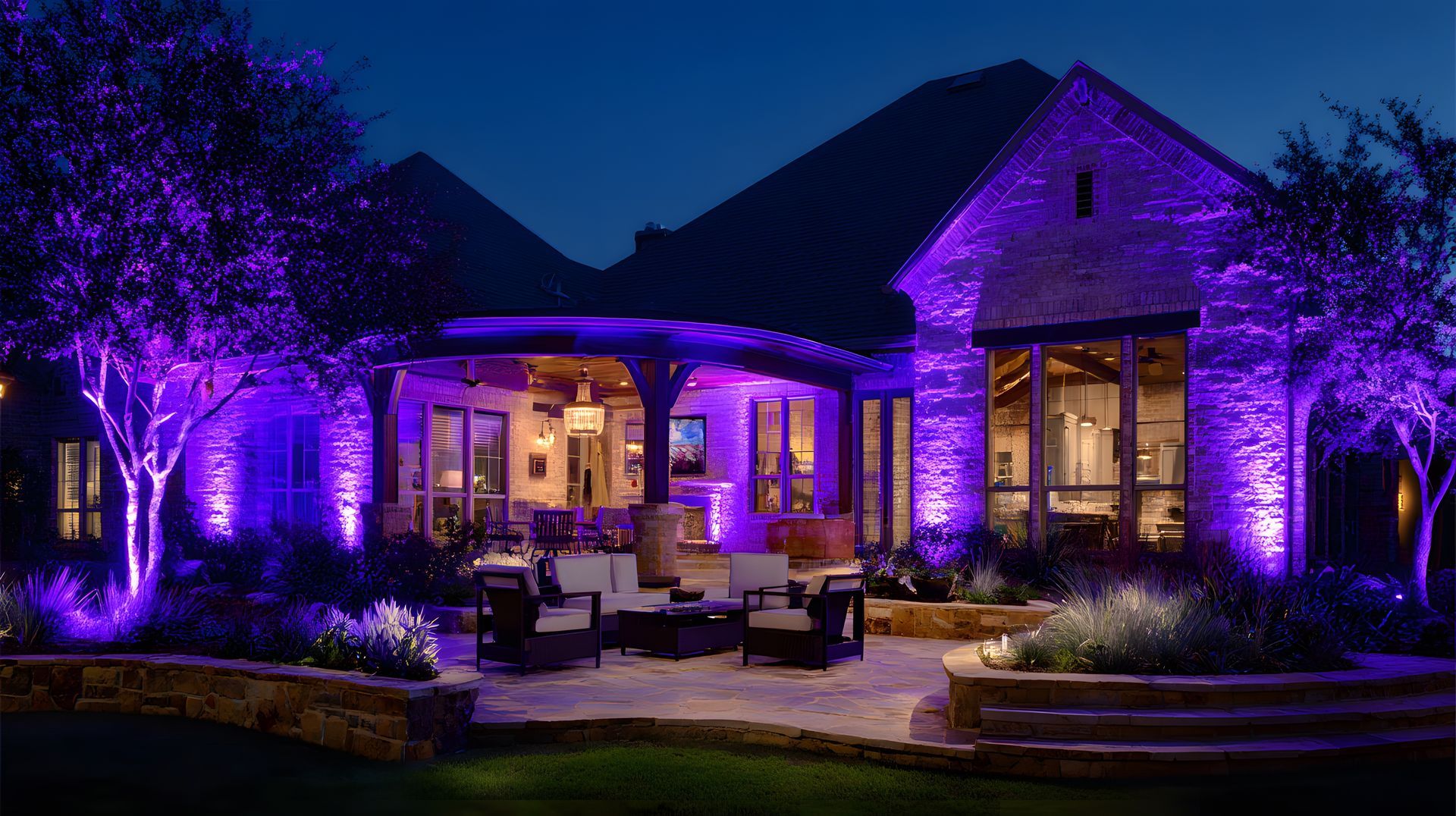 Purple-lit stone house with a warmly lit patio and outdoor seating at night