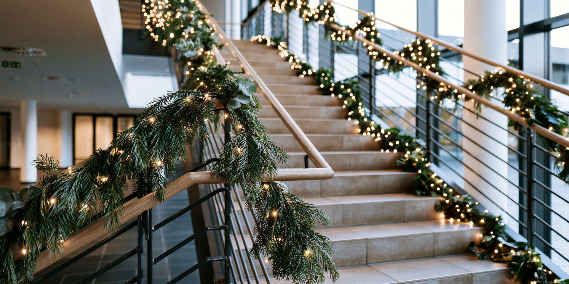 Indoor staircase decorated with evergreen garlands, warm lights, and white floral accents along the railing
