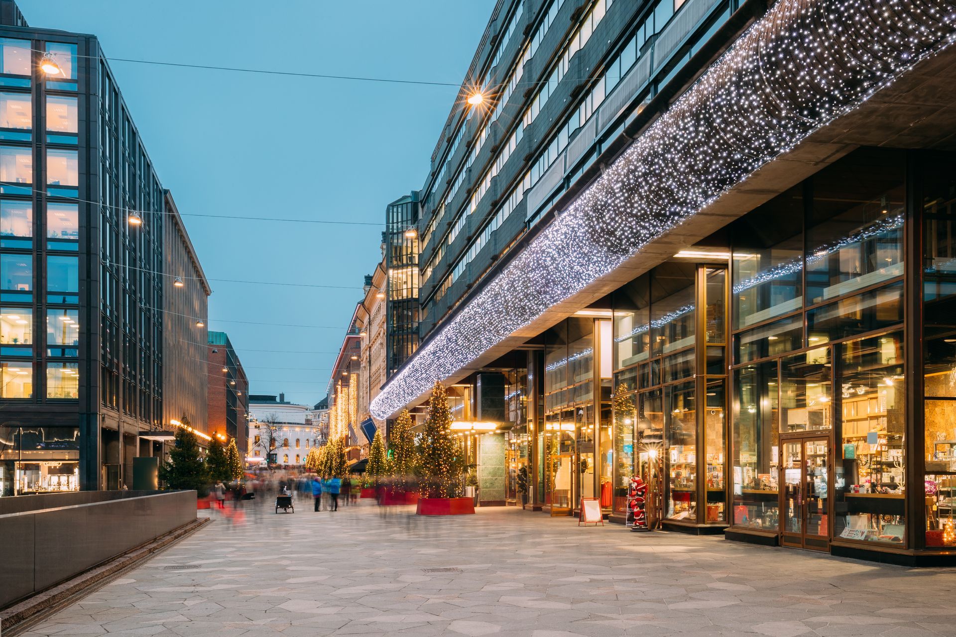 Modern shopping street at dusk with glass-fronted buildings, string lights, and pedestrians walking along the plaza