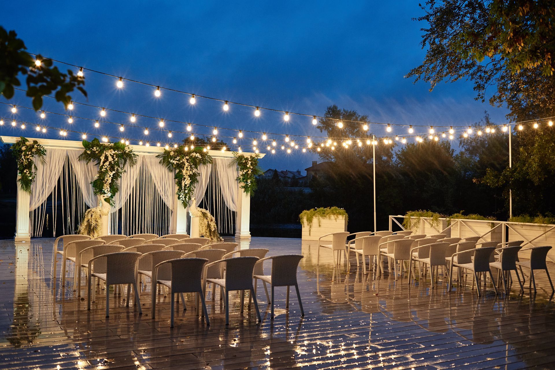 Wedding ceremony setup at dusk: white chairs face a decorated arch with string lights overhead.