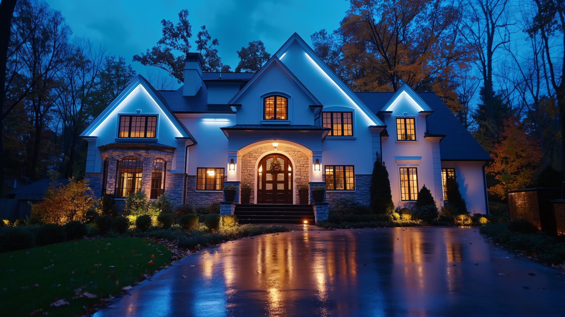Large illuminated house at dusk with glowing windows, wet driveway, and trees silhouetted against a blue sky