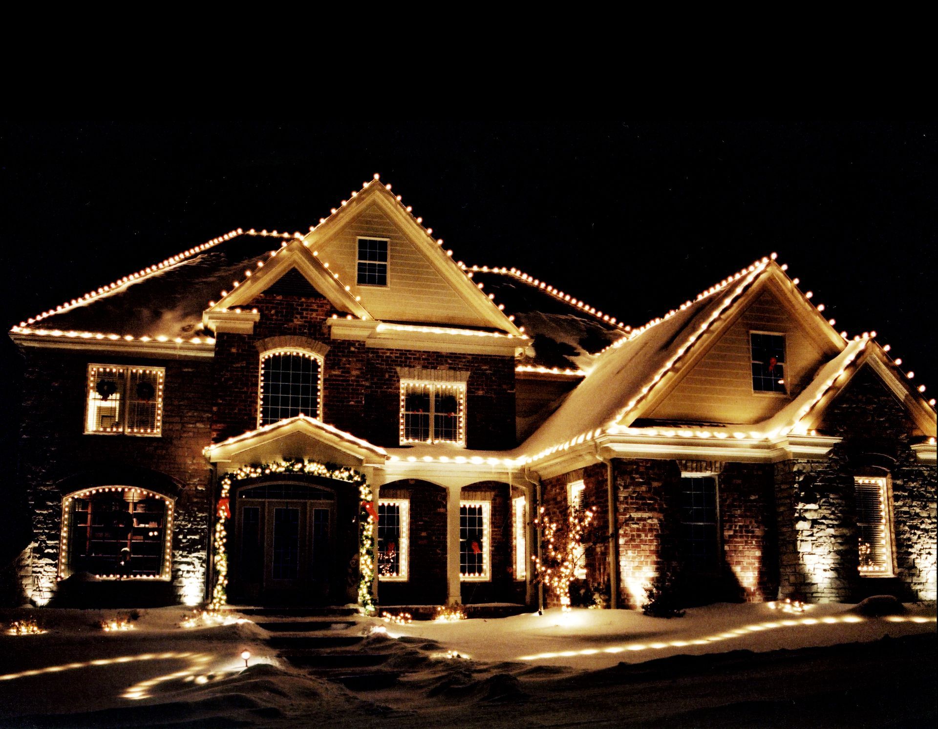 Large brick house illuminated with Christmas lights against a dark night sky, snow on the ground.