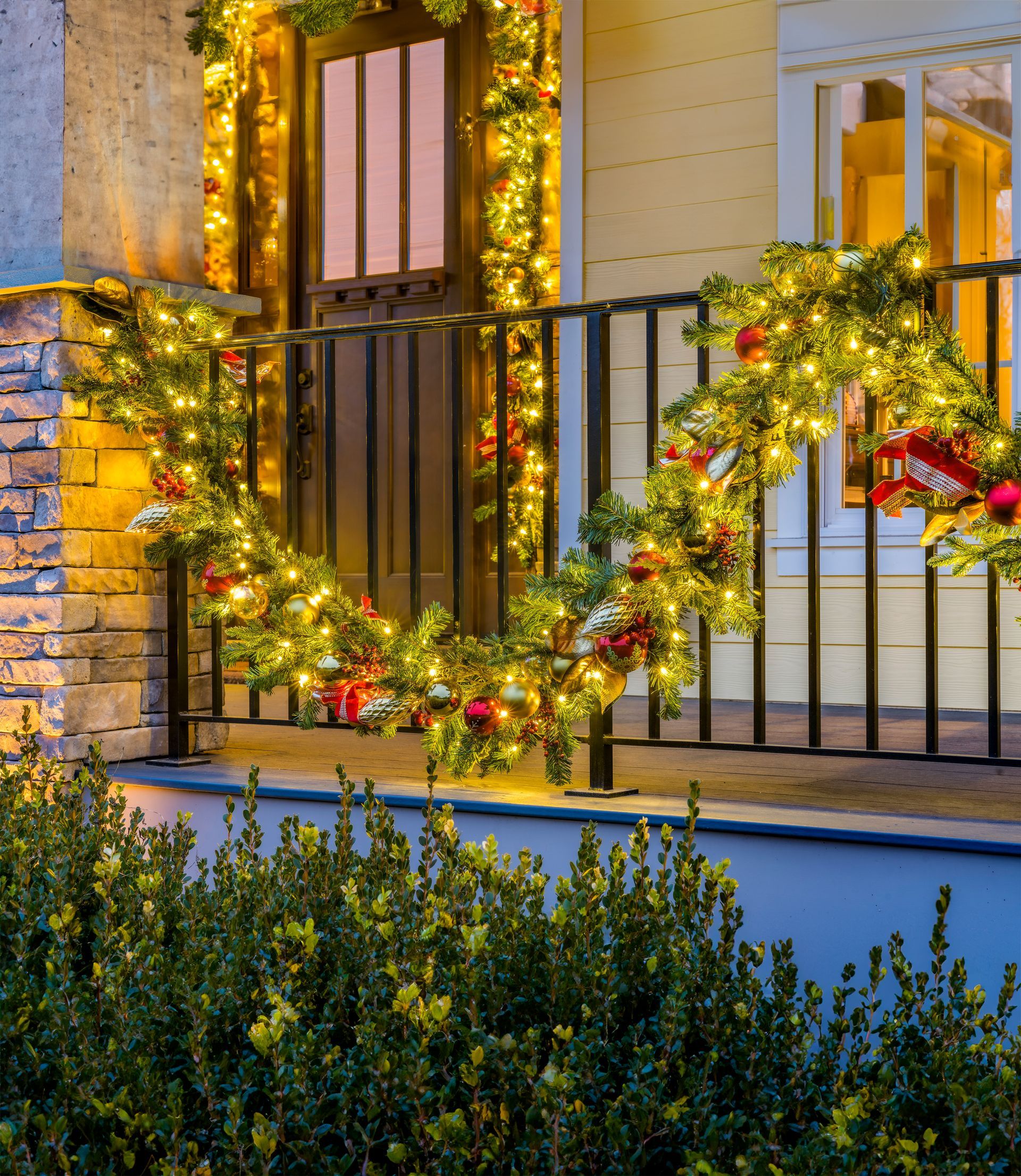 Christmas garland with lights and red ornaments on a porch railing near a door.