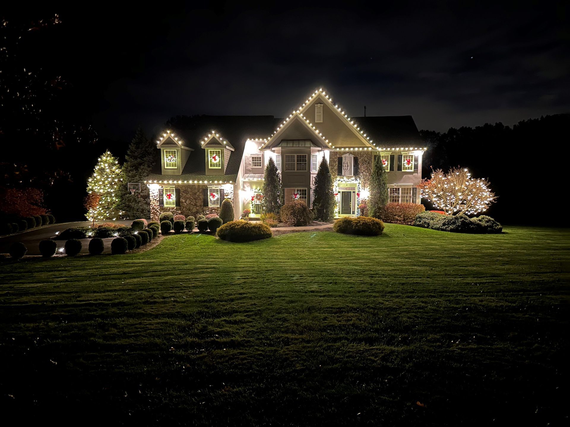 House at night, decorated with white Christmas lights. Green lawn, bushes, and trees add to the festive scene.