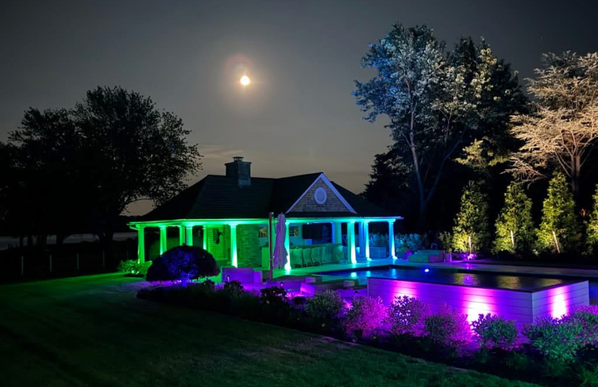 Night view of a house with colorful green, blue, and purple landscape lighting under a bright moon.
