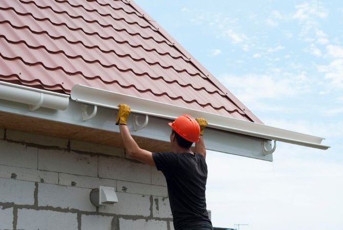 Worker in orange hard hat installing gutter beneath a red tiled roof