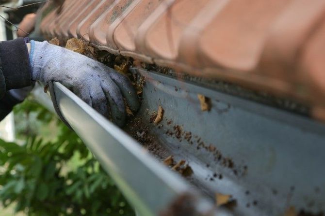 Gloved hand cleaning dirt from a gutter beneath terracotta roof tiles