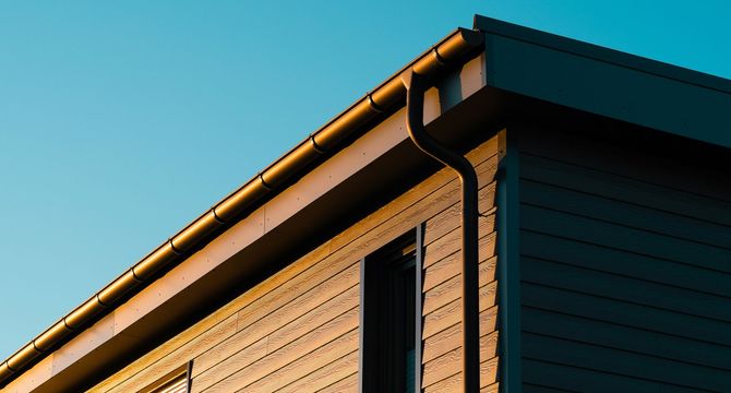 Close-up of a house corner with tan siding, dark trim, and a teal roof against a blue sky