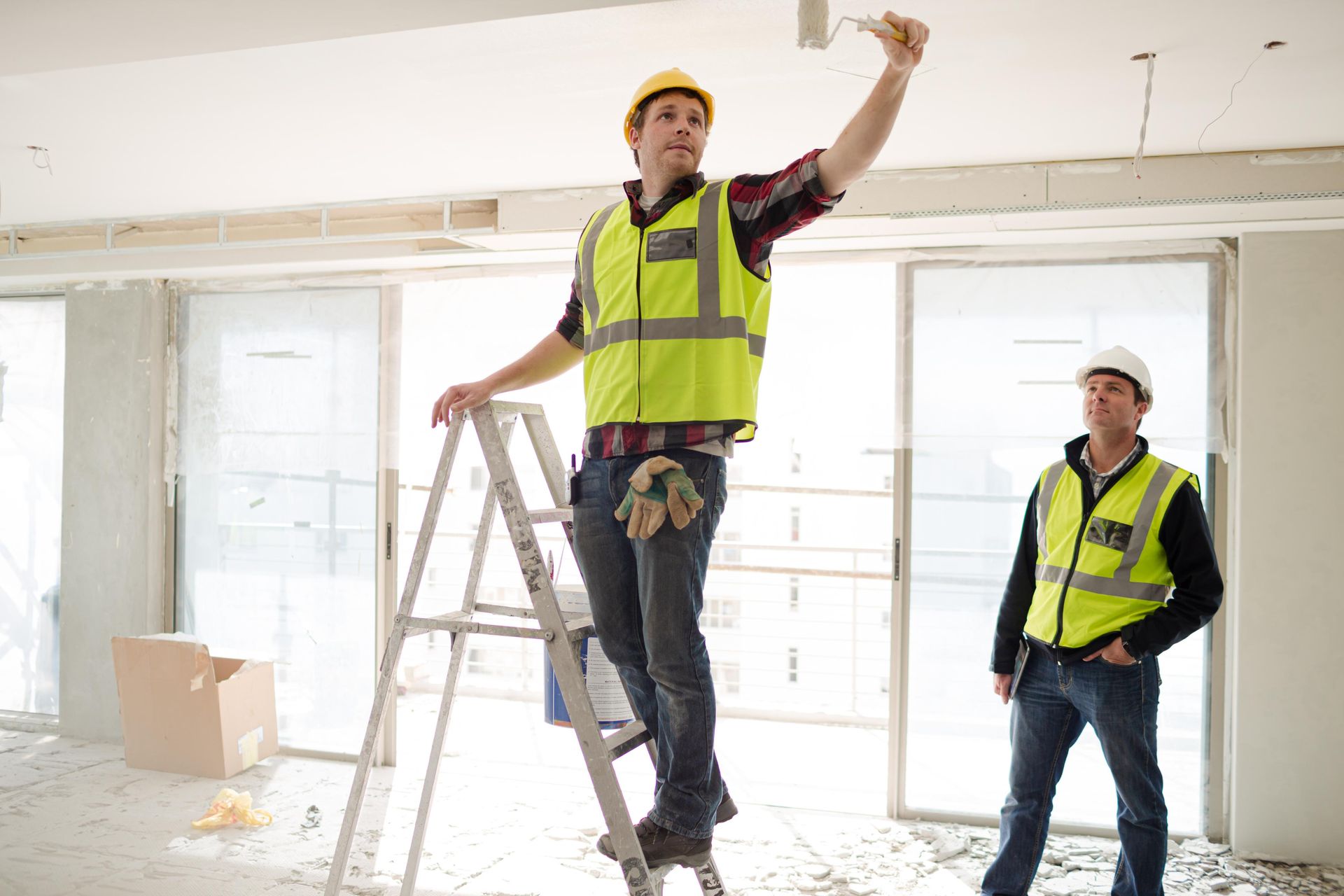 Two construction workers painting a ceiling, one on a ladder, wearing safety vests and hard hats.