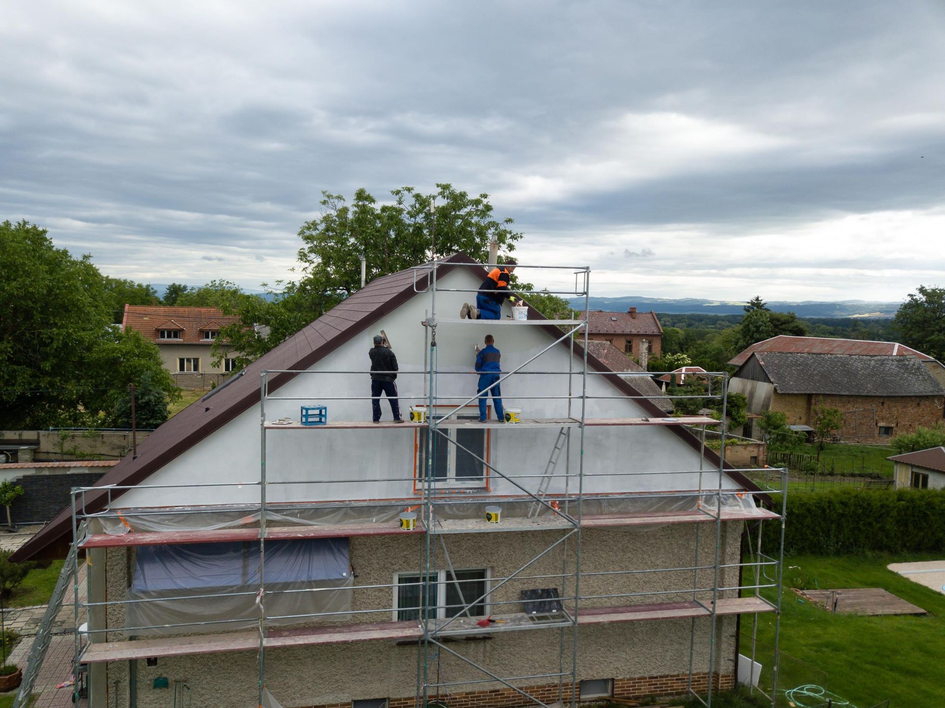 Workers on scaffolding repairing a house exterior. The sky is cloudy, the roof is brown, and the house is white.