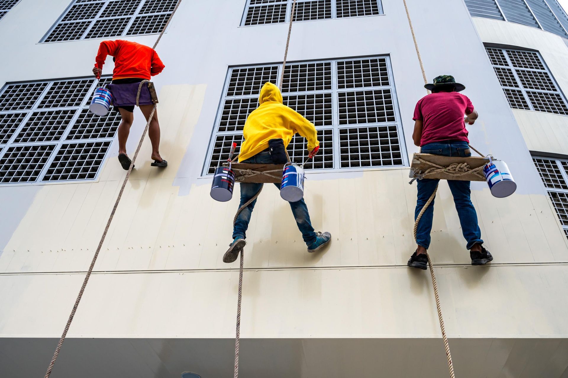 Three painters on ropes paint a white building. The workers wear colored clothes and have paint buckets.
