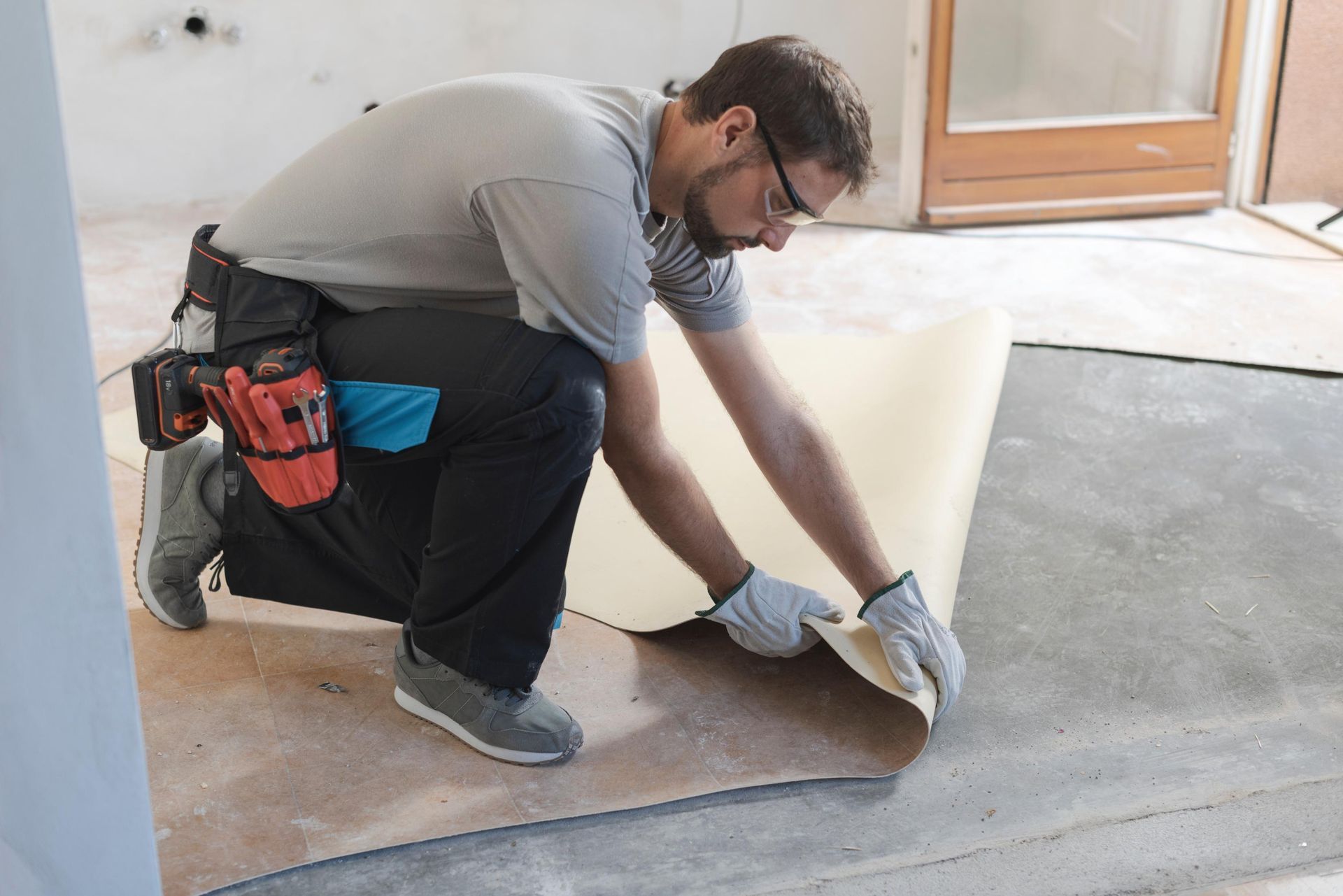 Man kneeling, installing flooring, peeling up sheet. Gray shirt, black pants, tool belt, gloves.