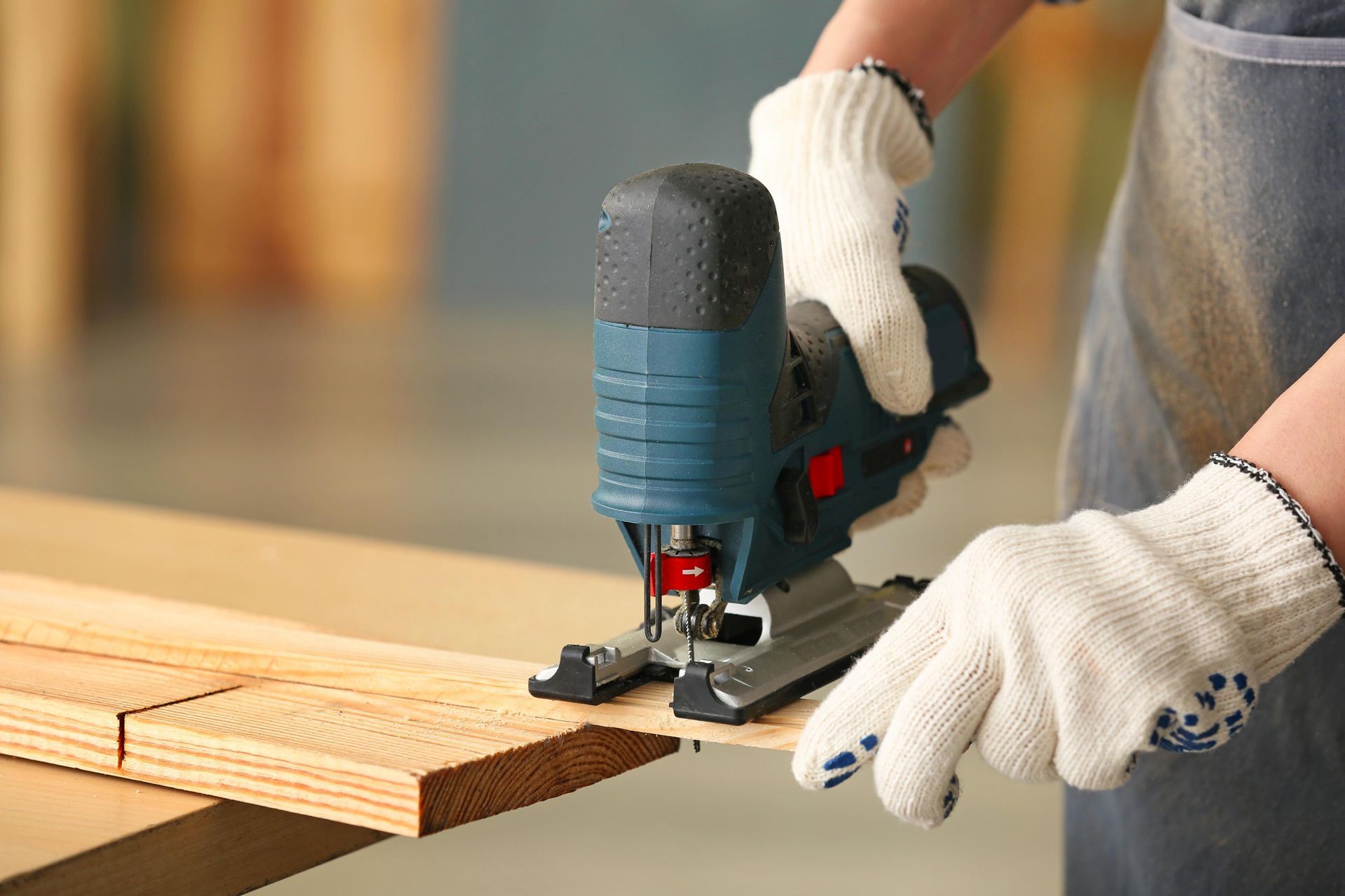 Person using a jigsaw to cut wood, wearing gloves, indoors.