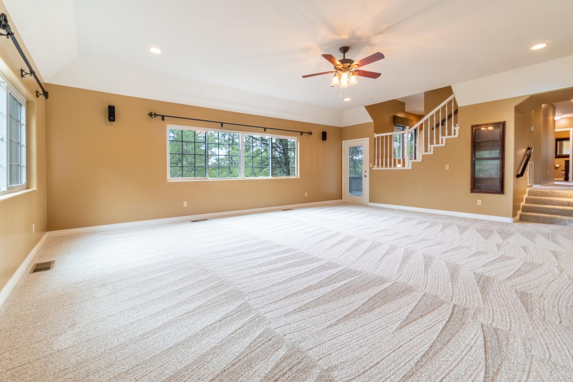 Spacious, empty room with tan walls, white carpet, and stairs leading to a second level.