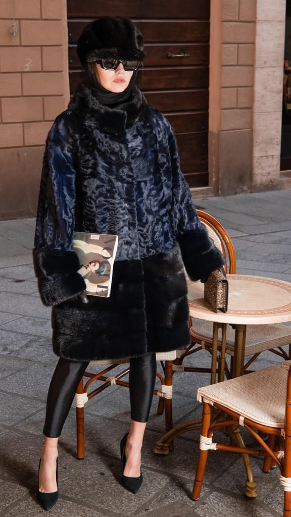 Donna con pelliccia scura, cappello, occhiali da sole e leggings, con in mano una pochette mimetica, seduta accanto a un tavolo all'aperto.