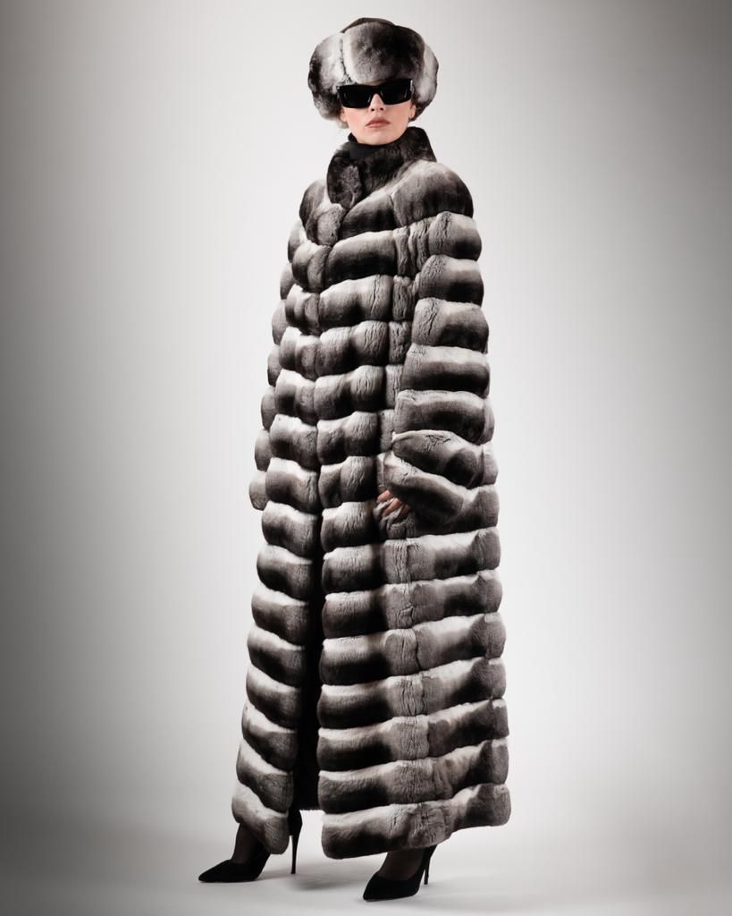 Donna con pelliccia lunga fino ai piedi, cappello e occhiali da sole, in piedi su uno sfondo grigio.