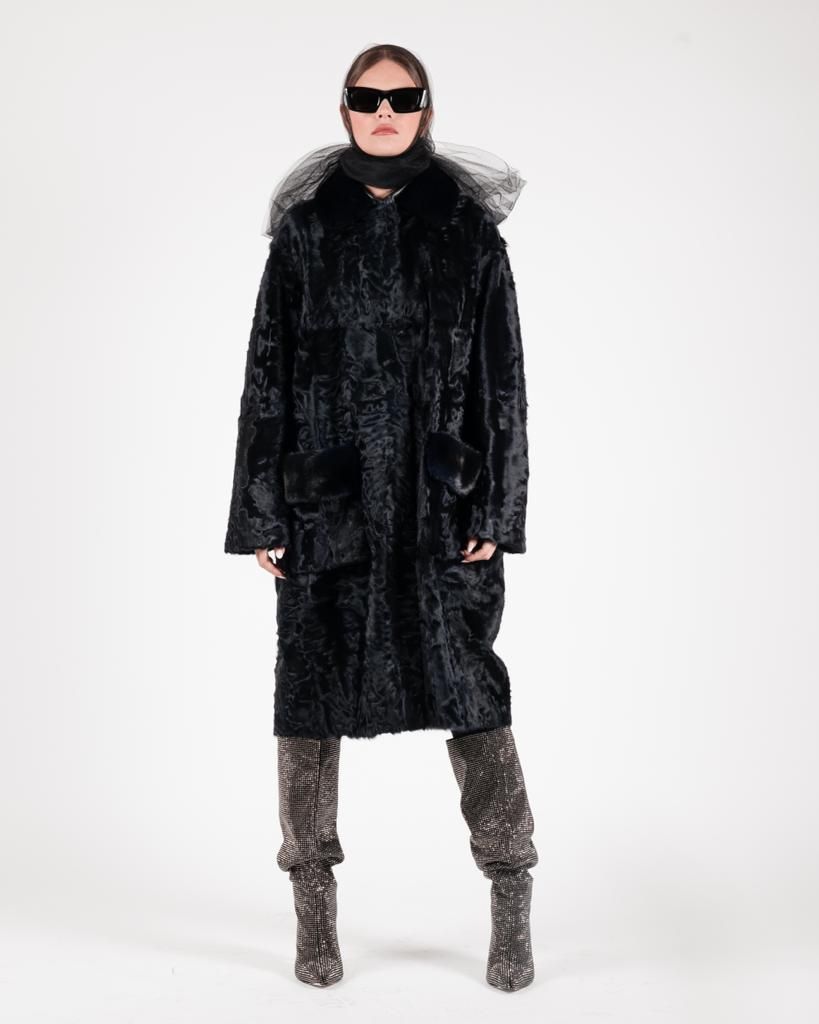 Donna che indossa un lungo cappotto nero, occhiali da sole e stivali, in posa davanti a uno sfondo bianco.