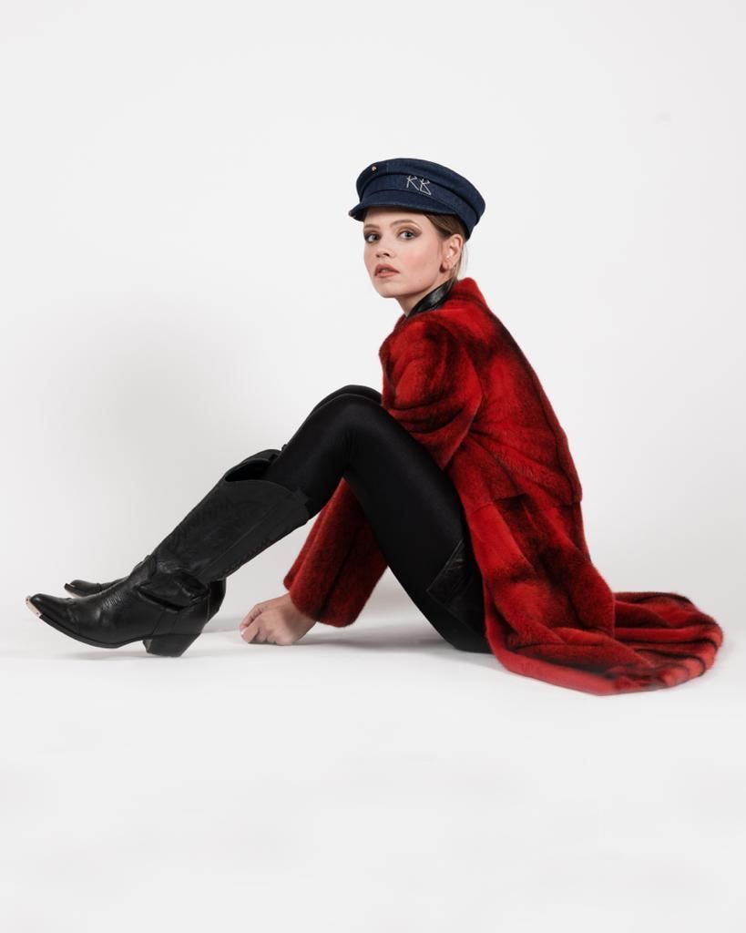 Una donna con pelliccia rossa, leggings neri e stivali è seduta su uno sfondo bianco, indossa un cappello blu scuro e guarda oltre la spalla.