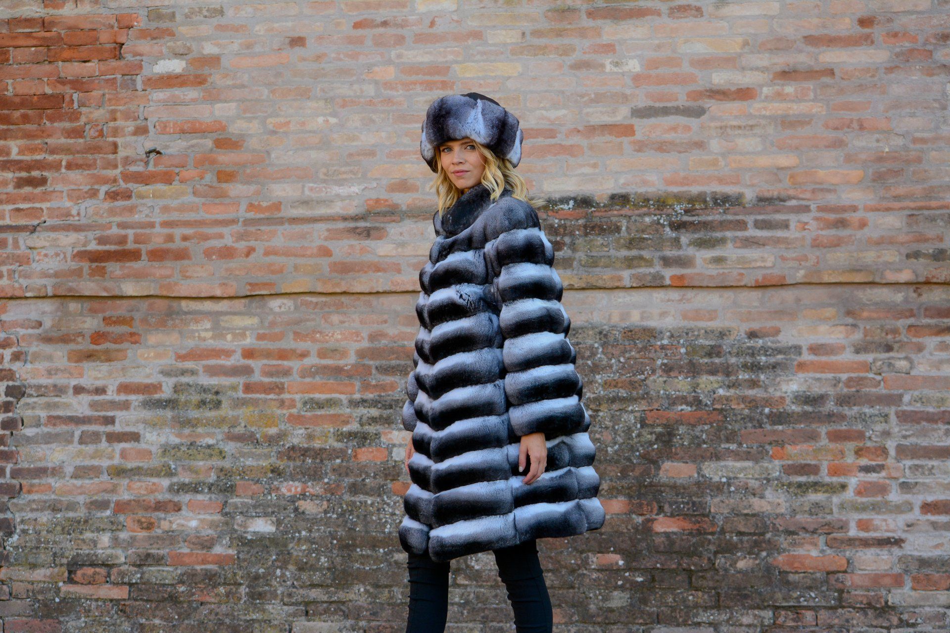 Una donna con pelliccia e cappello è in piedi davanti a un muro di mattoni. Il cappotto è a strisce bianche e nere.