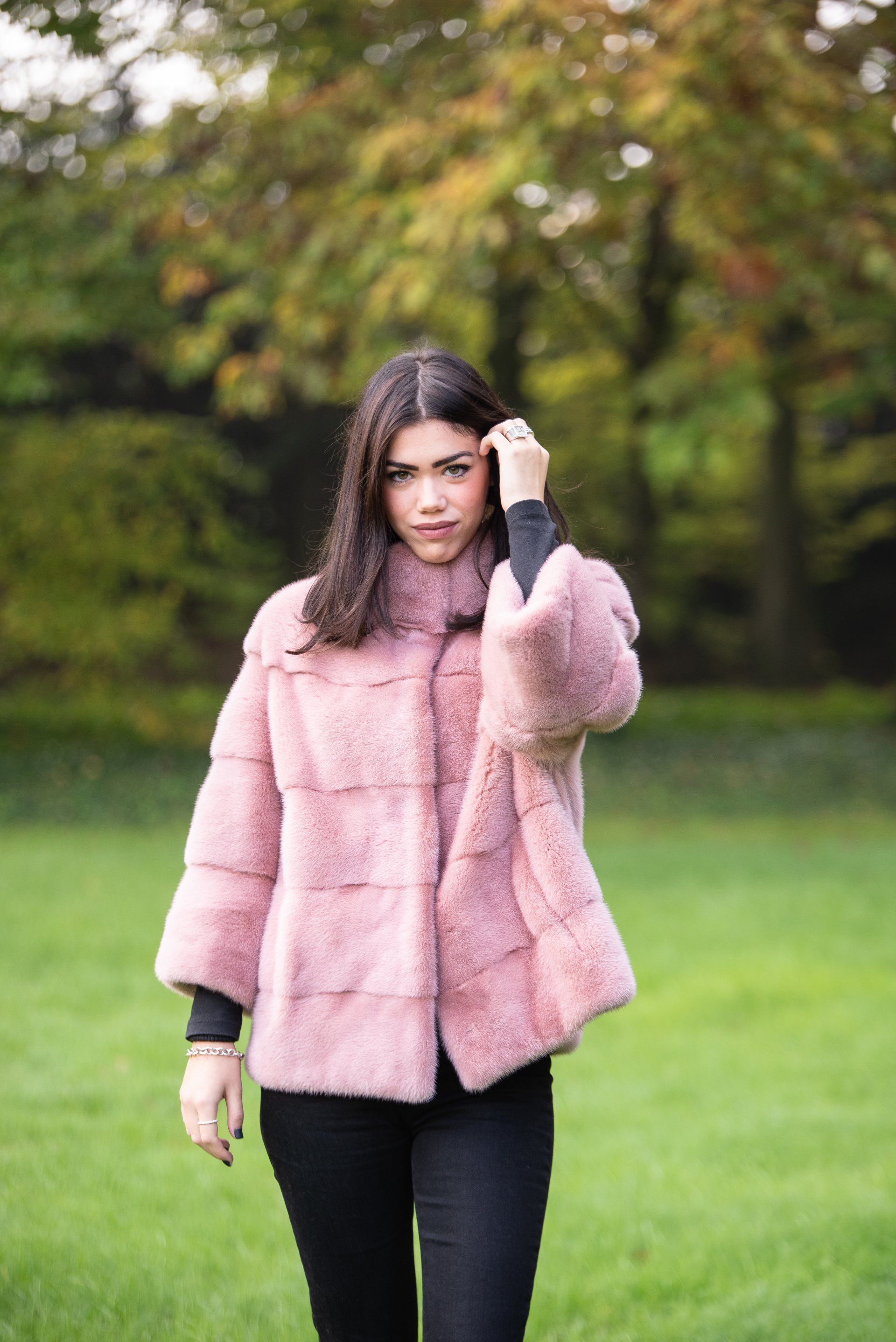 Donna con pelliccia rosa, top e pantaloni neri, in piedi in un'area erbosa con alberi.