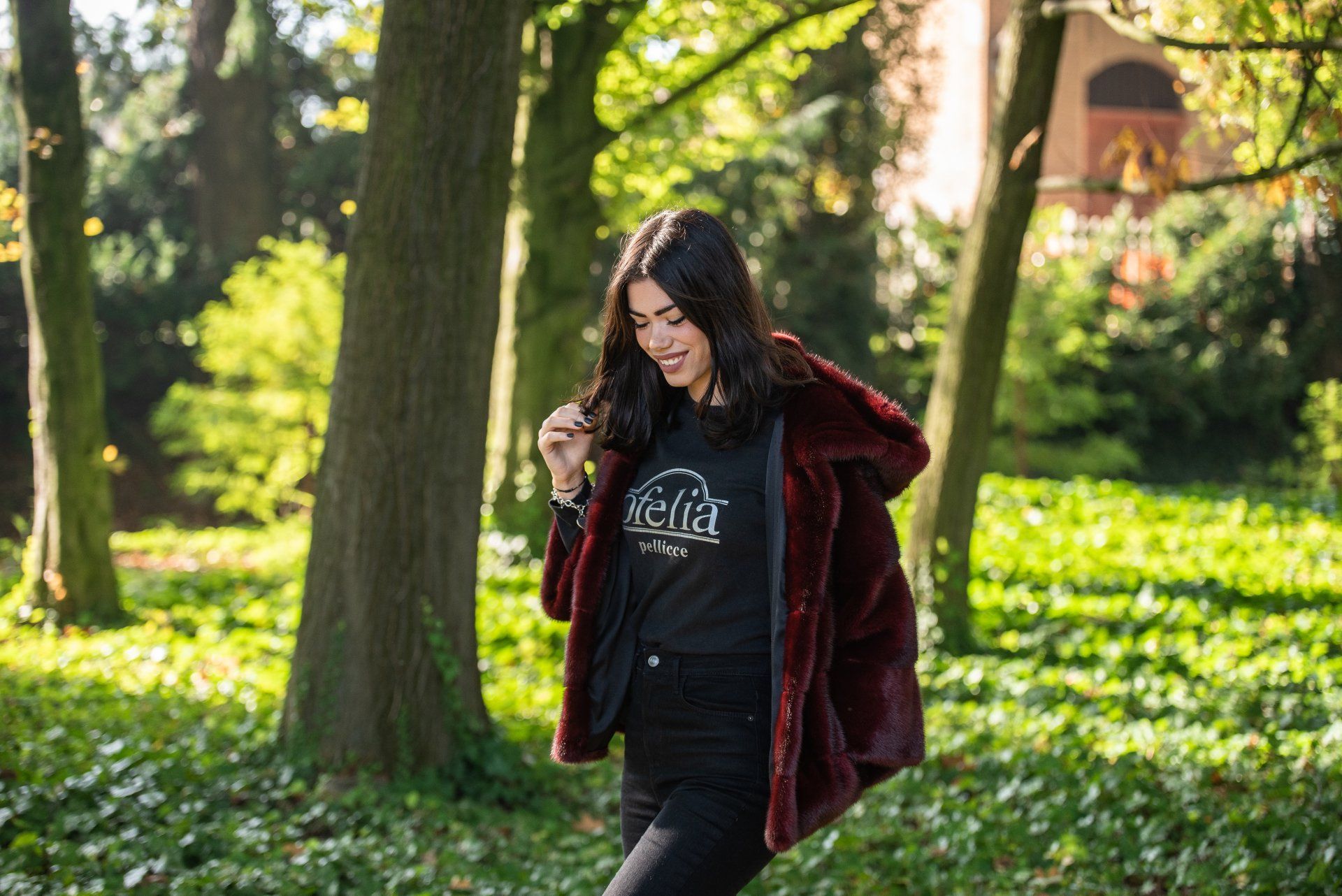 Donna sorridente con un cappotto di pelliccia sintetica color bordeaux, in piedi in un parco soleggiato.