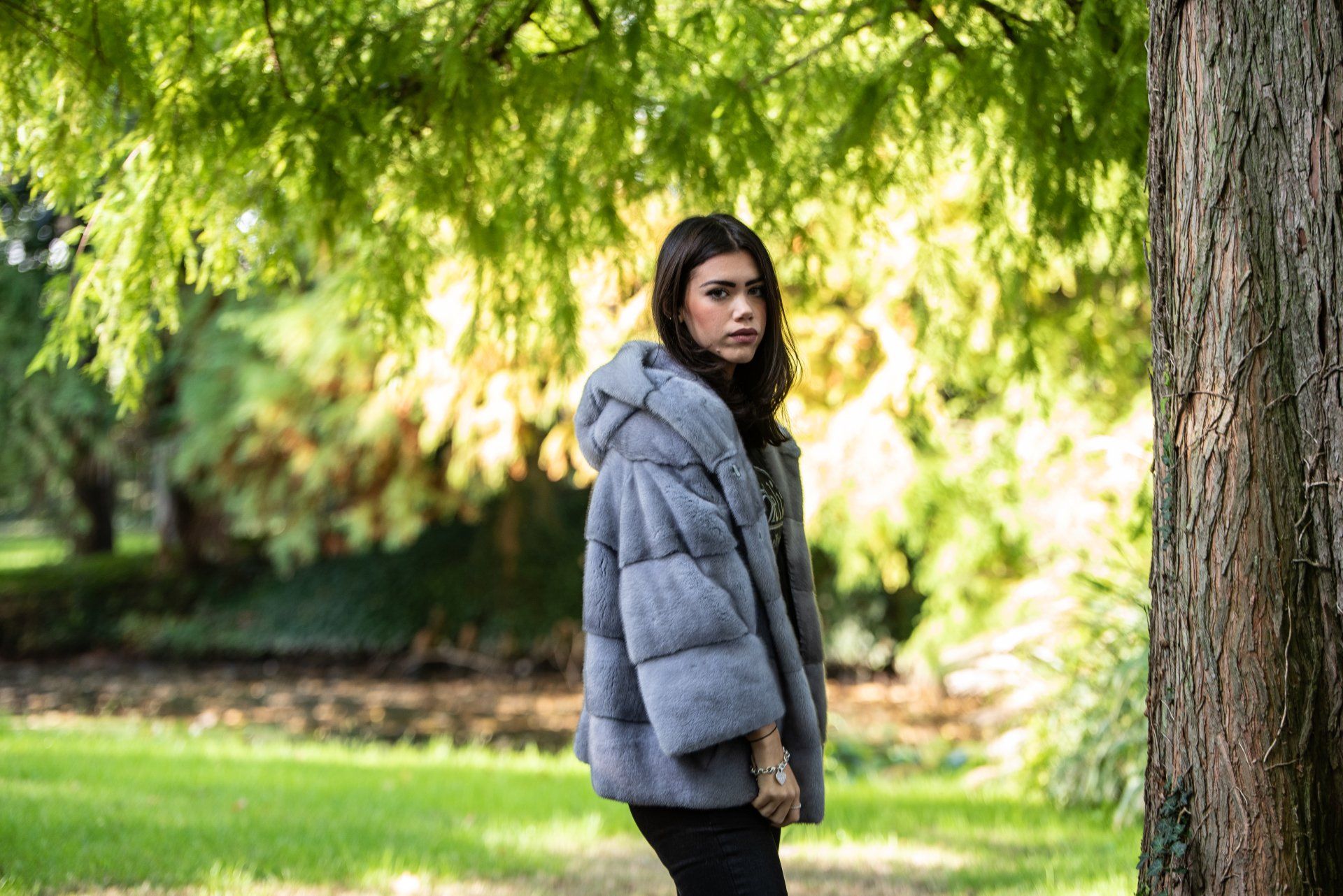 Una donna con un cappotto di pelliccia grigia con cappuccio è in piedi vicino a un albero, in un parco con erba verde e fogliame.