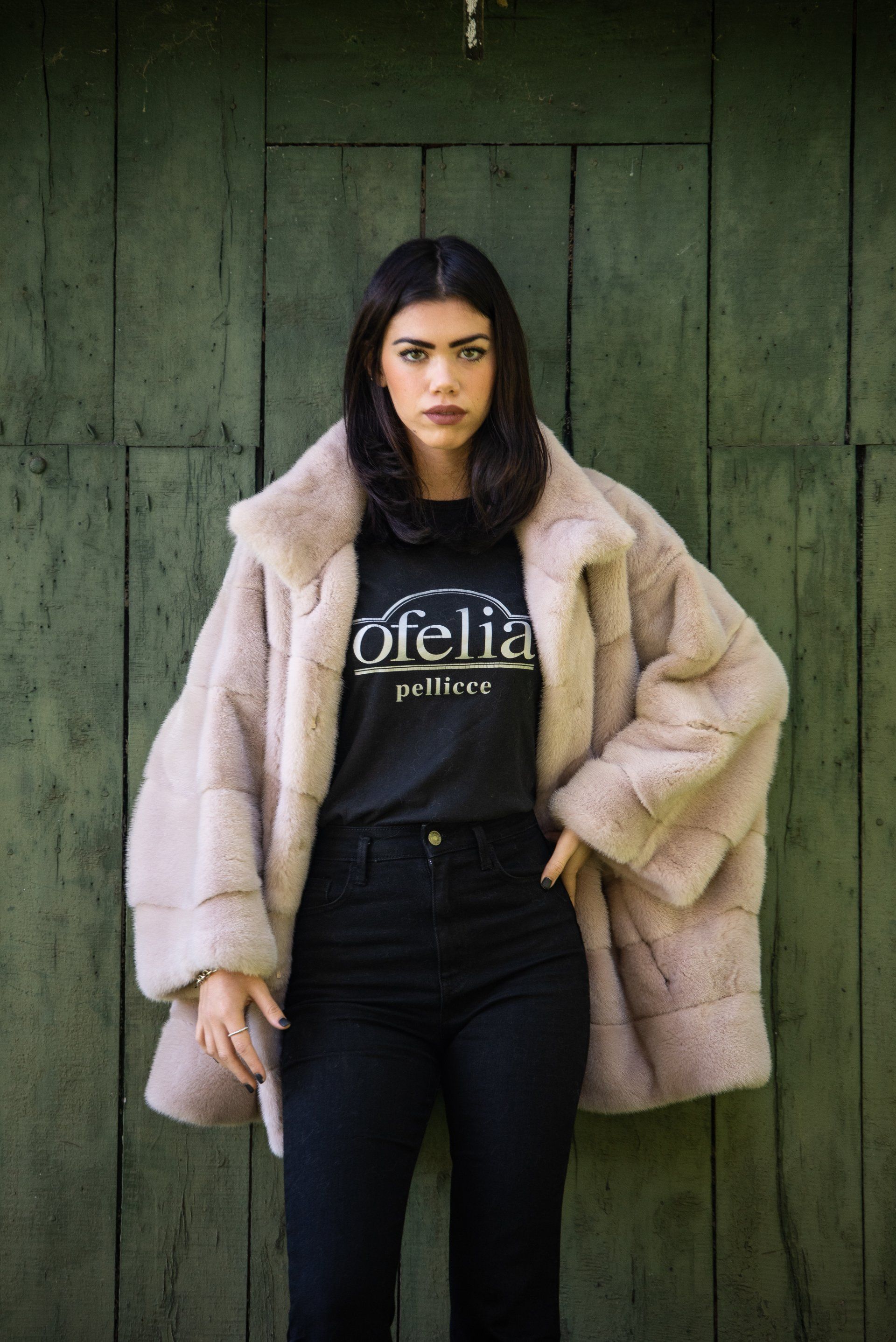 Donna che indossa un cappotto di pelliccia sintetica color marrone chiaro e jeans neri, in piedi contro una porta di legno verde.