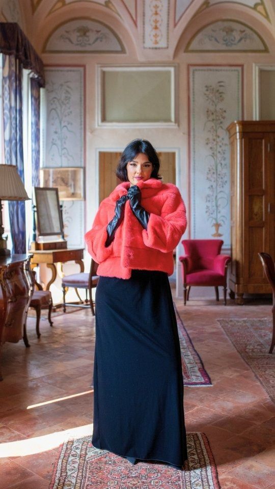 Una donna con un cappotto rosso e un abito nero, e guanti neri, è in piedi in una stanza con dipinti murali decorati.