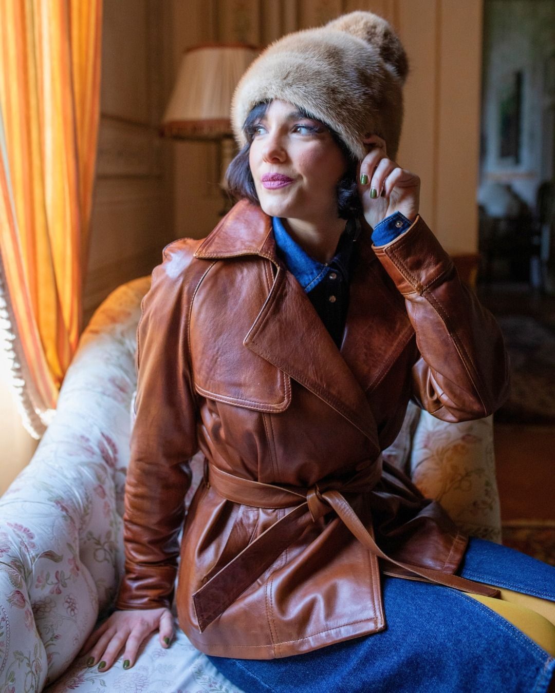 Donna con giacca di pelle marrone e cappello di pelliccia, seduta all'interno, che guarda altrove.
