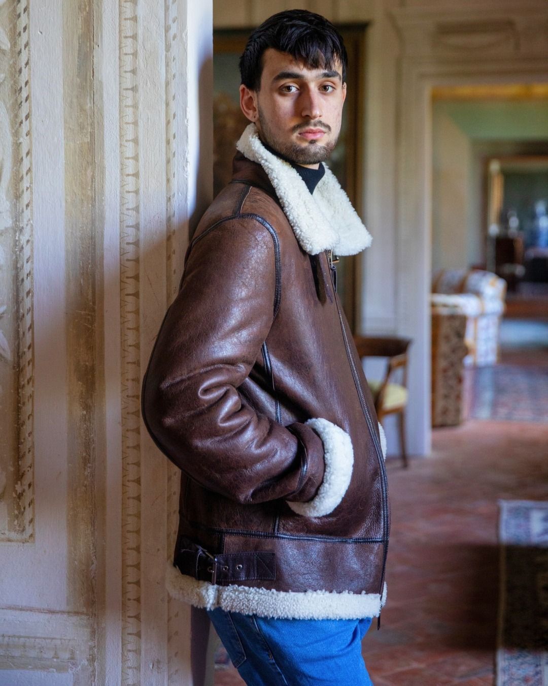 Uomo con giacca di montone marrone e jeans blu. Appoggiato a un muro in casa.