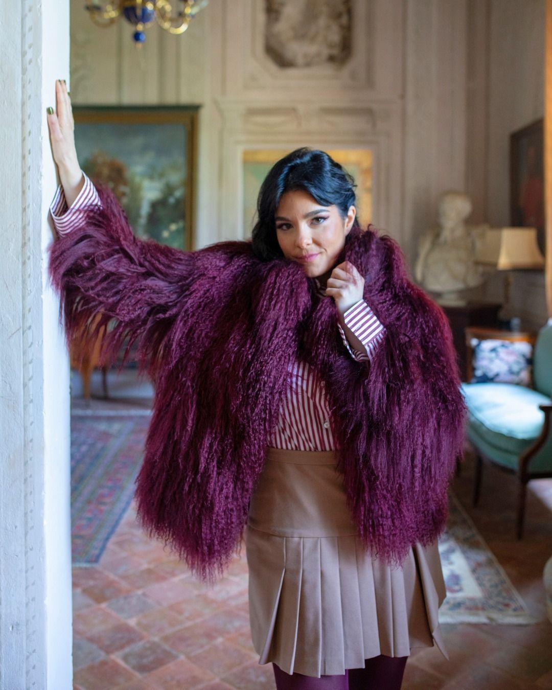 Donna con giacca di pelliccia sintetica viola e gonna a pieghe, appoggiata a un muro in una stanza con opere d'arte e decorazioni elaborate.