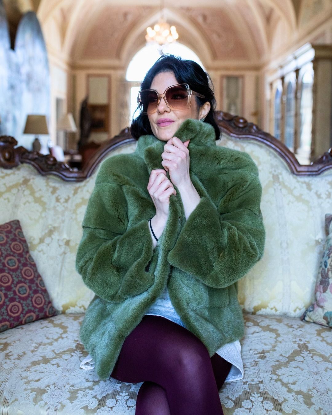Donna con pelliccia verde, occhiali da sole e leggings color castagna, seduta su un divano d'epoca in una stanza con porte ad arco.